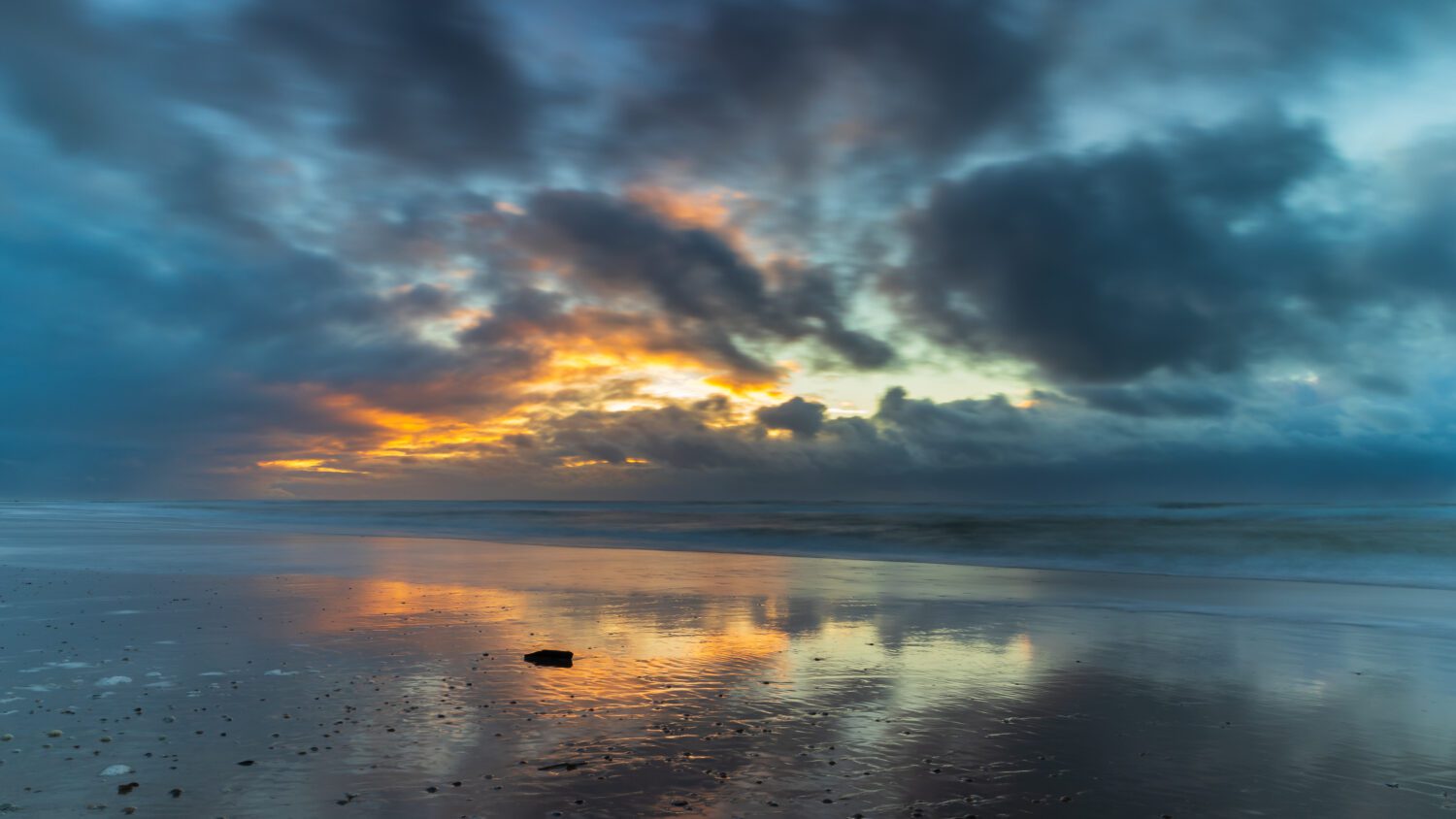 Een stuk hout in de branding van de Noordzee waardij de ondergaande zon en dreigende wolken vechten om voorrang