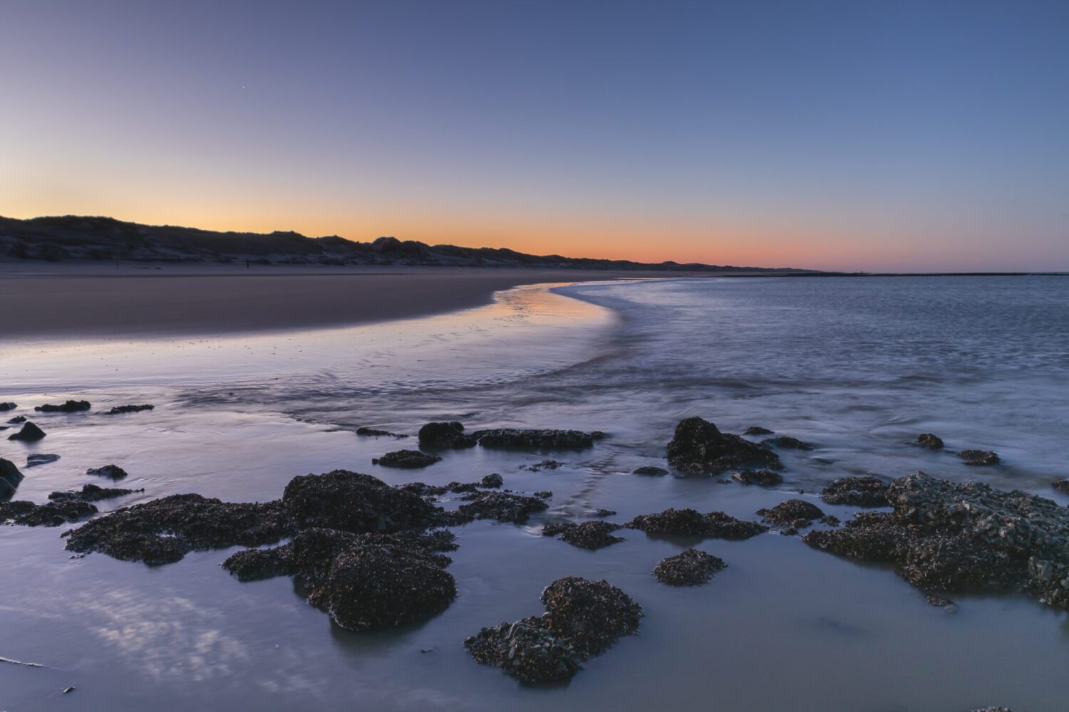 Vanaf de pier leidt de golvende kustlijn van de Noordzee naar de warme kleuren van de opkomende zon