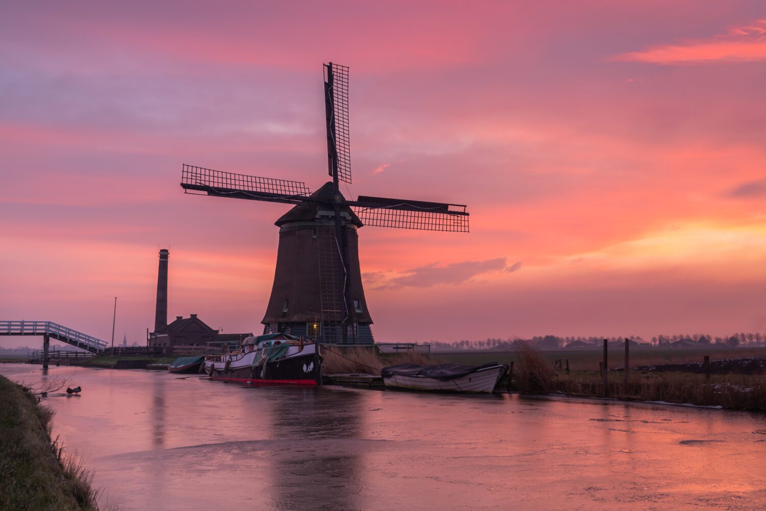 Een roze lucht en bevroren sloot met boten vormen het decor voor de Kaagmolen en naastgelegen boogbrug en stoomgemaal