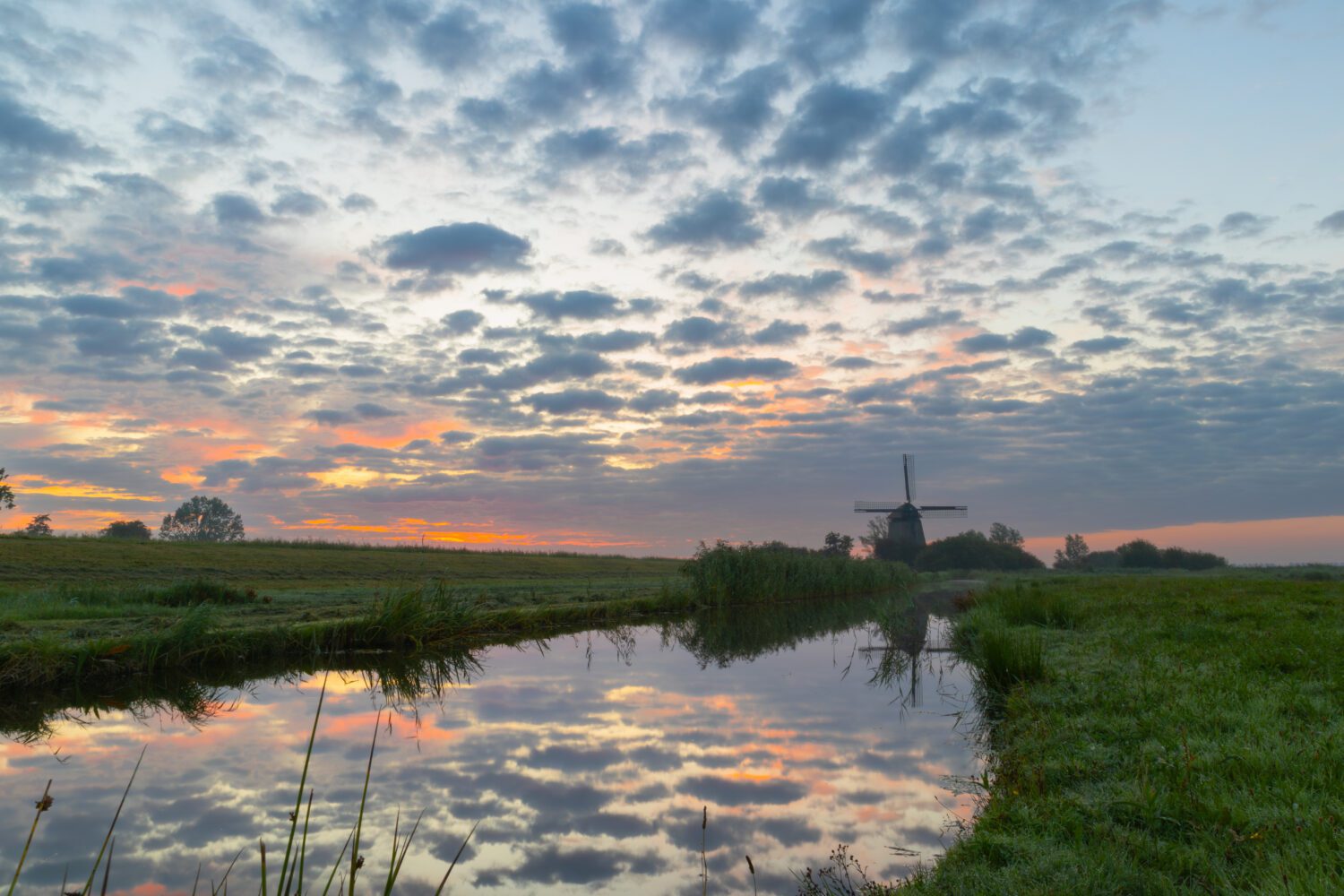 De rode lucht bedekt met plukjes wolken reflecteren in de sloot naast molen De Havik in Grootschermer