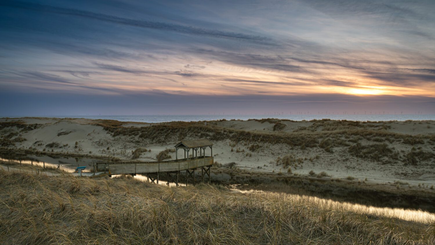 Een overdekte houten steiger tussen de duinen bij Petten met zicht op de ondergaande zon boven de Noordzee
