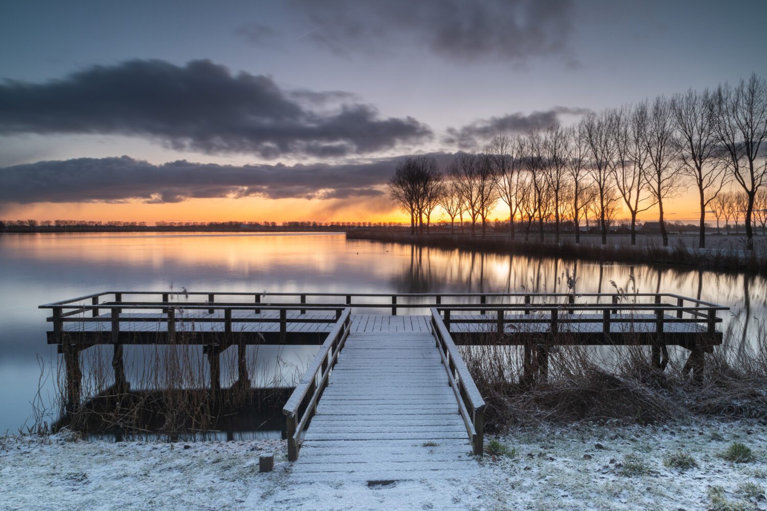 De opkomende zon schijnt door de bomen naast het Meer van Dirkshorn waar de steiger bedekt is met sneeuw