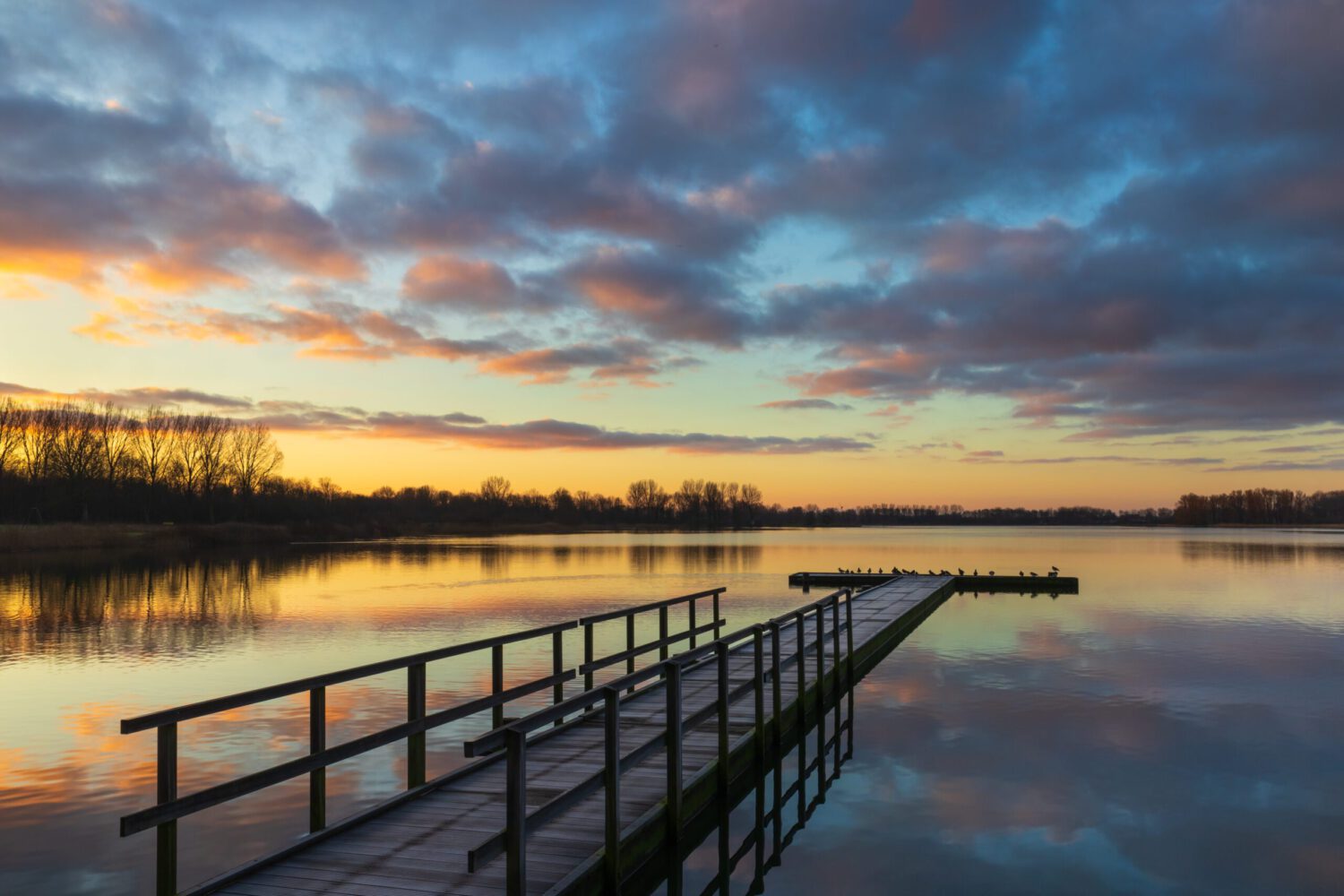Een kleurrijke wolkenlucht tijdens zonsopkomst boven de steiger in het meer van recreatiegebied Geestmerambacht