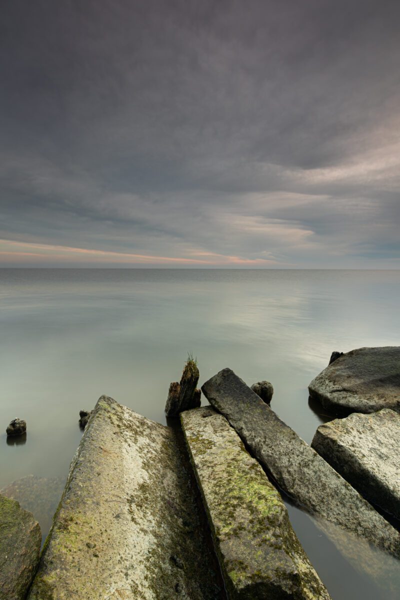 Stenen en houten palen met gras in het rustige water van het Markermeer tijdens een bewolkte zonsopkomst