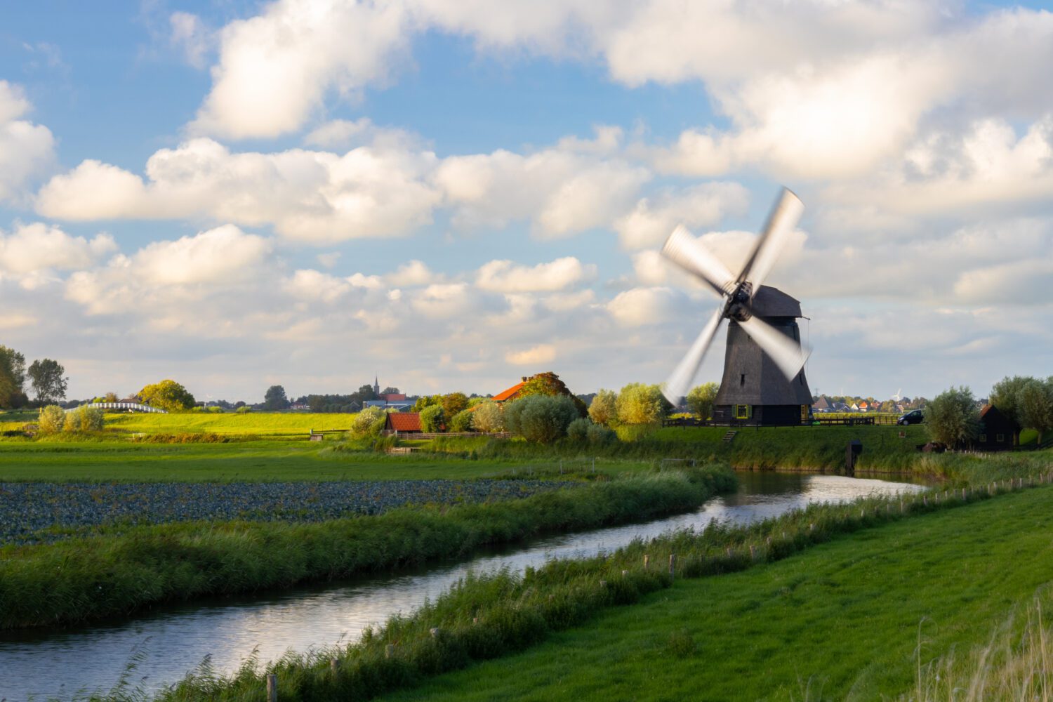 Een molen met draaiende wieken met wit doek aan de dijk onder een typisch Hollandse blauwe lucht met witte wolken