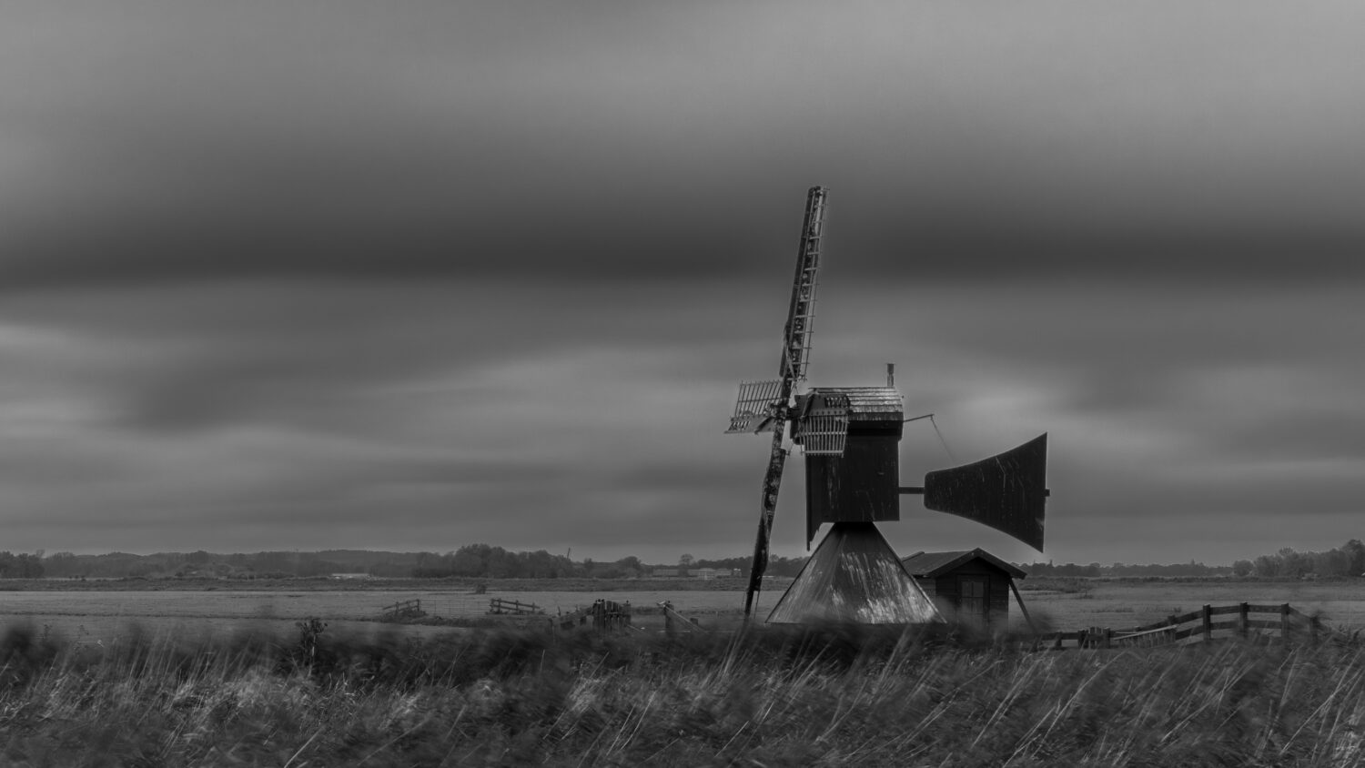 Zwartwit foto van de Varnebroekermolen in het weiland bij Heiloo onder donkere wolken en omgeven door riet en houten hekken