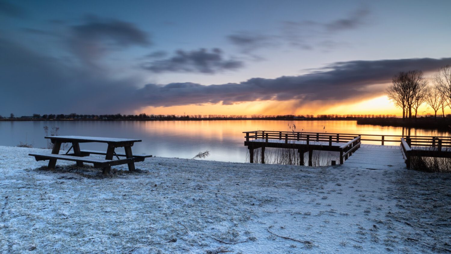 Het meer van Dirkshorn in winterse setting met sneeuw op de picknicktafel en steiger en op de achtergrond een winterse bui voor de opkomende zon