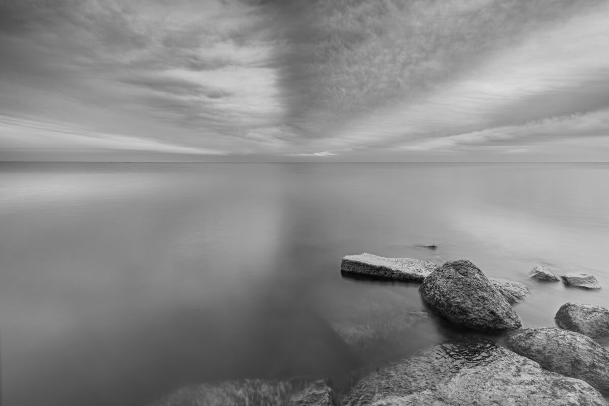 Zwartwit foto van stenen in het Markermeer onder een gestreept wolkendek
