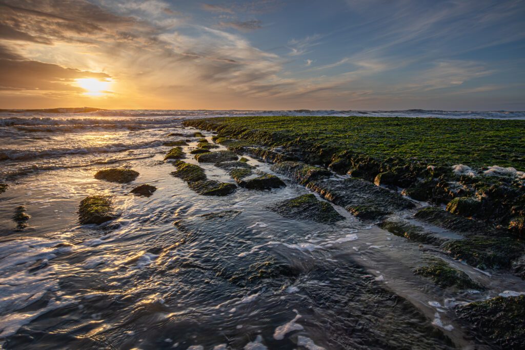 De ondergaande zon boven de Noordzee schijnt nog net door de wolken heen en verlicht de met zeewier bedekte pier en reflecteert in het rustig kabbelende water over de stenen naast de pier.