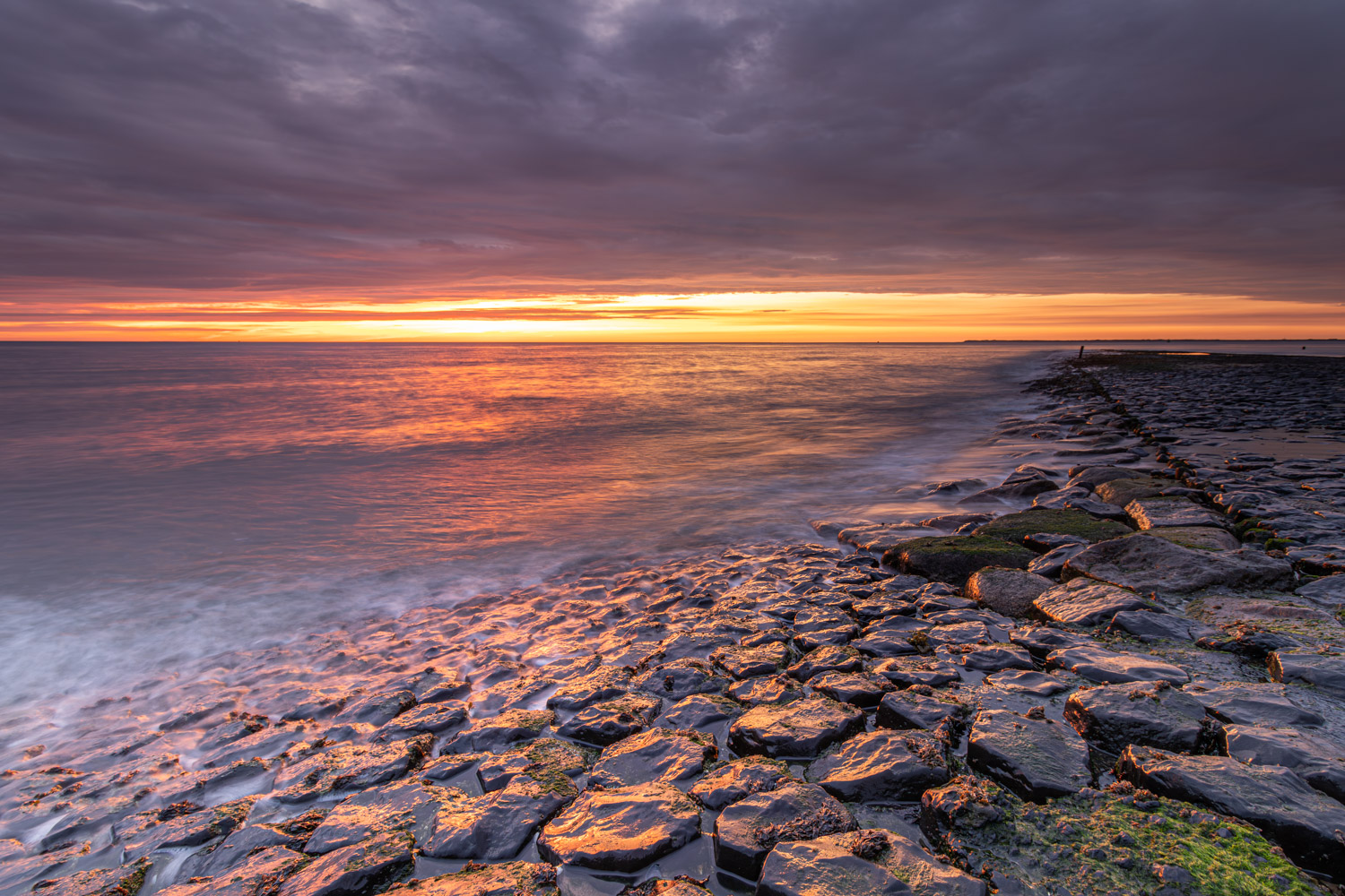 De zeepier in de Noordzee wordt zachtjes overspoelt met water, terwijl de ondergaande zon weerspiegelt in het zeewater en de dreigende wolken doet kleuren.