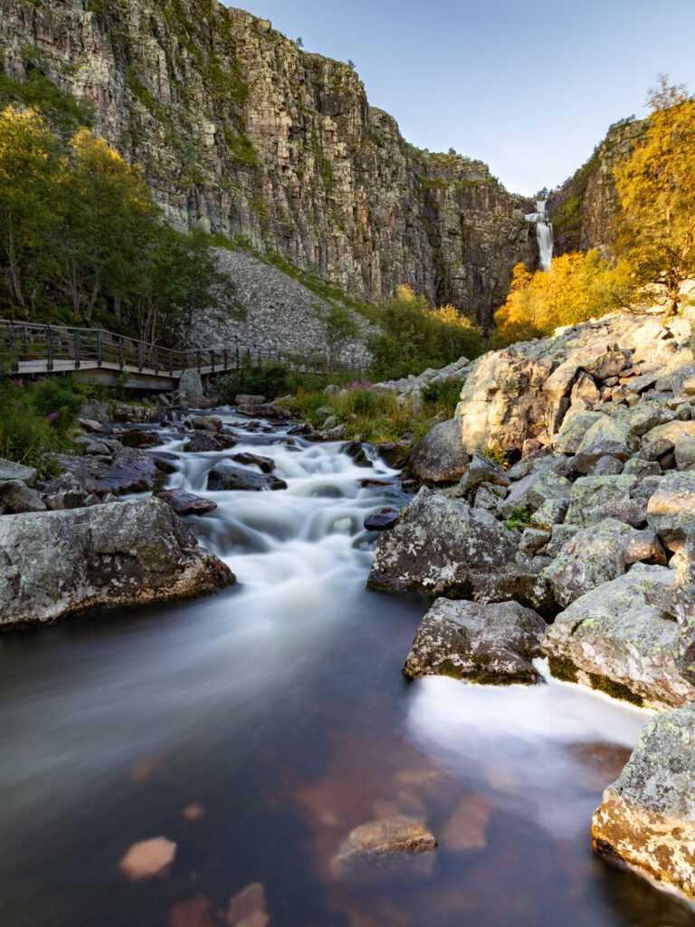 De Njupeskärs waterval in Zweden wordt verlicht door de opkomende zon, waarbij op de voorgrond het stromende water voorbij spoelt.