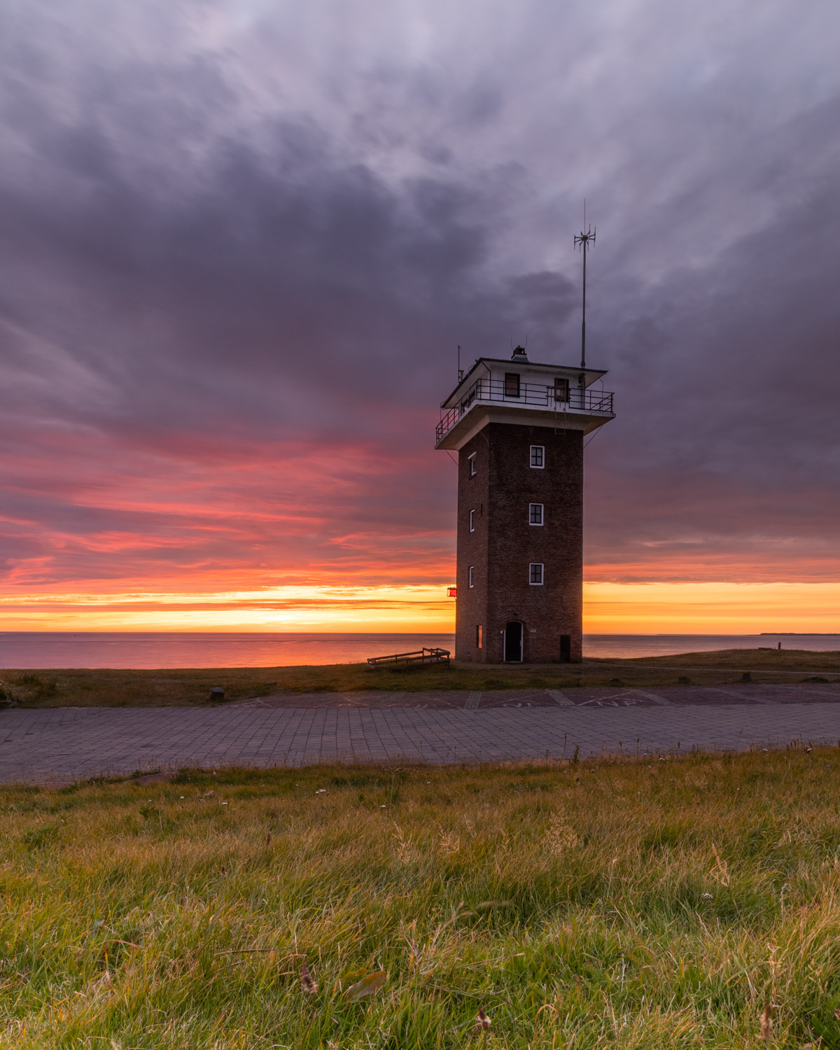 De oude vuurtoren op de dijk aan de Noordzee onder een donkere dreigende wolkenlucht, terwijl de ondergaande zon zorgt voor prachtige oranje en rode kleuren.