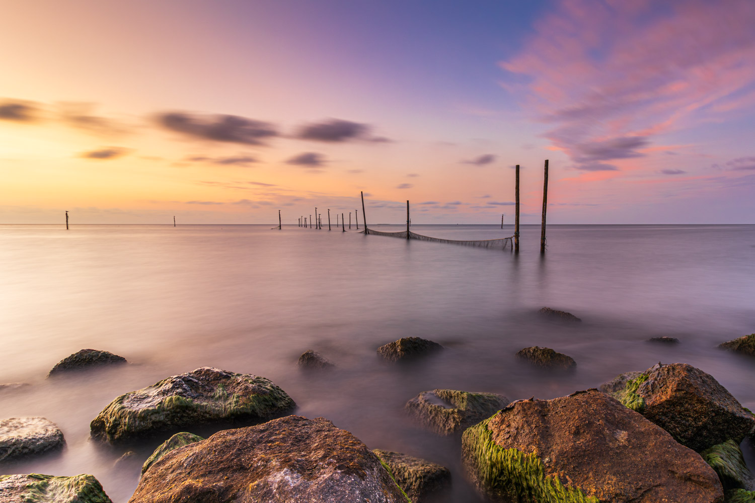 Onder een kleurrijke lucht tijdens zonsondergang steken de palen met visnetten en rotsen langs de kant nog net boven het water van het IJsselmeer uit.
