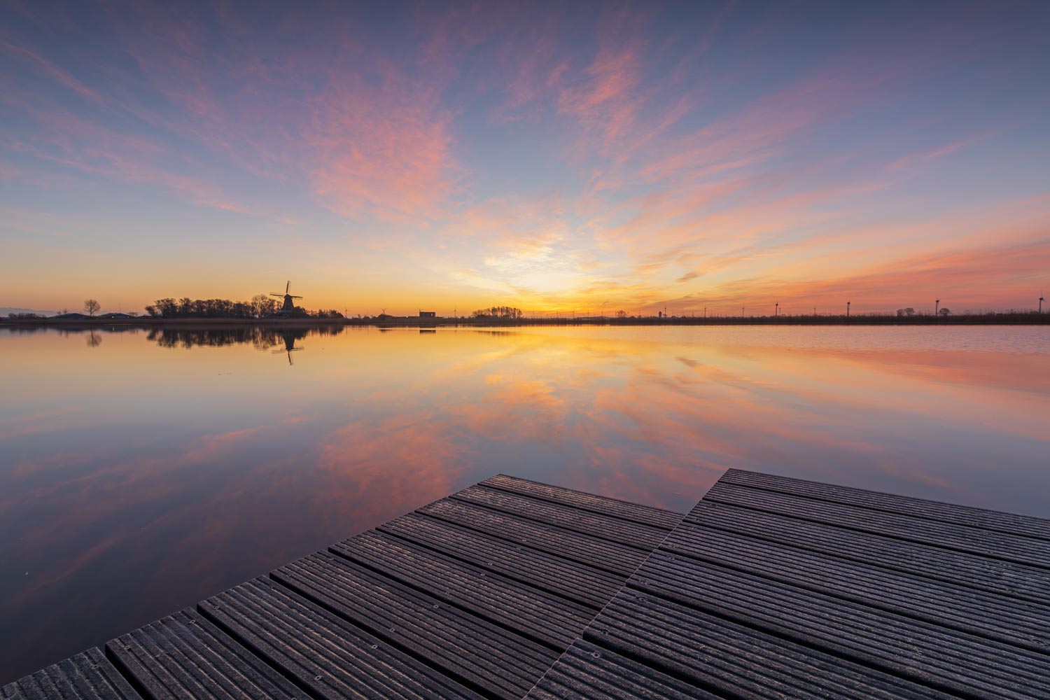 De hoeken van de houten steiger wijzen naar de molen aan de overkant van het spiegelgladde water met de reflectie van de kleurrijke wolken tijdens zonsopkomst