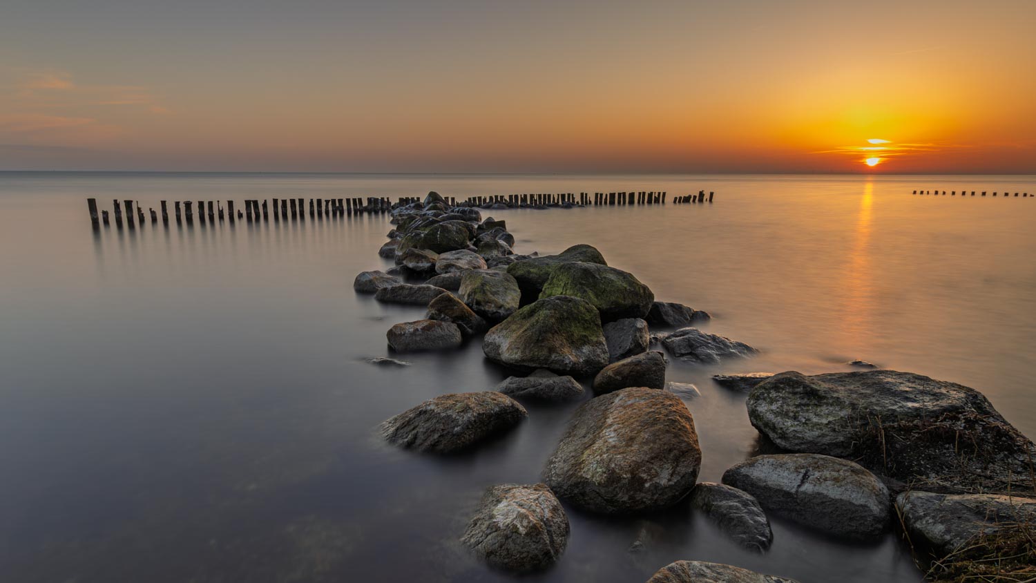 Golfbreker van basalt blokken en houten palen in het IJsselmeer leiden naar de gouden zo die net boven de horizon uitkomt